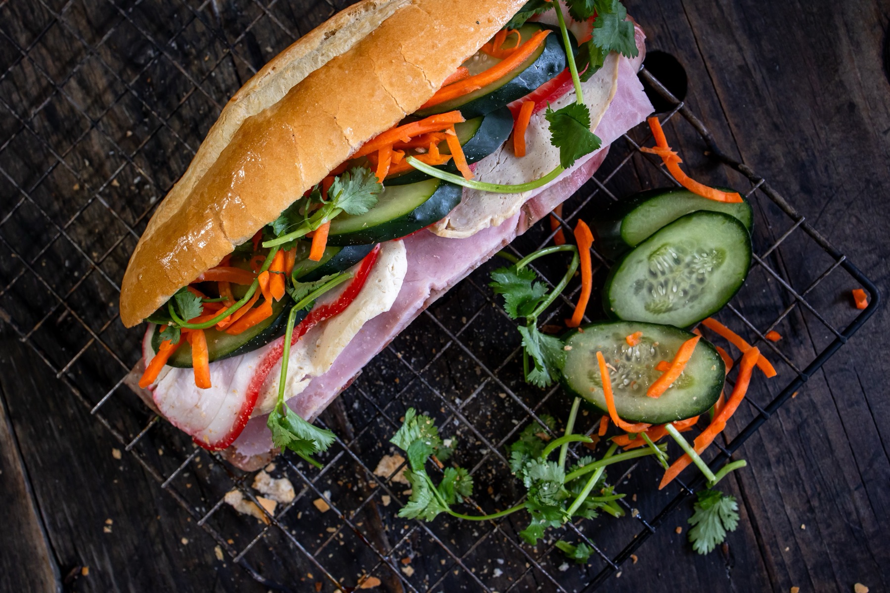 Classic combination bánh mì resting on a wire rack, topped with cucumber, pickled carrot, and cilantro